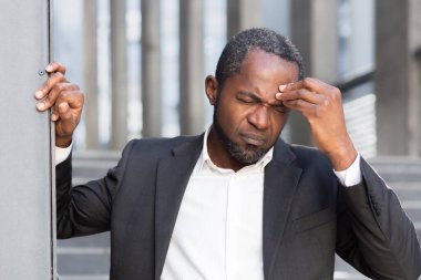 Senior African American man suffering from severe headache, pressure. He is standing outside the office center, leaning against the wall, holding his head, grimacing in pain.