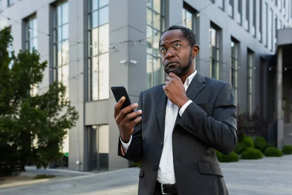 A serious African American businessman in a business suit is standing on the street near a skyscraper wearing a suit and glasses. He holds the phone in his hands, looks thoughtfully to the side.
