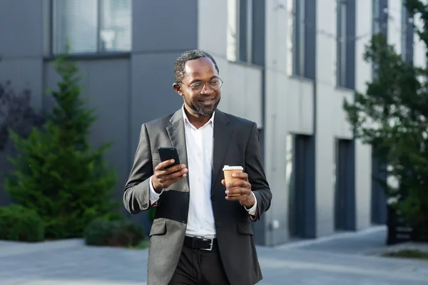 Coffee break. A successful African American businessman in a business suit is standing on the street near a skyscraper in a suit and glasses, holding a cup of coffee to go and using a mobile phone.
