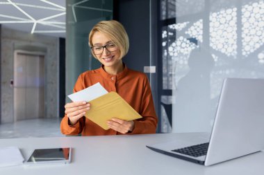 Successful business woman boss working inside office with laptop, employee received mail envelope letter with notification message, blonde woman reading and smiling good news, happy with achievement.