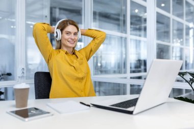 Portrait of happy female programmer inside office, female worker with laptop and headphones smiling and looking at camera, businesswoman satisfied with work and achievement results at workplace.