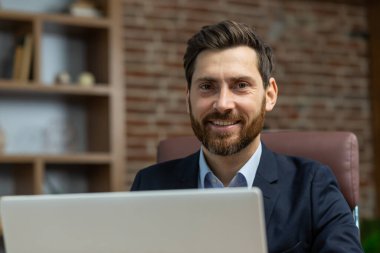 Ofis içinde başarılı yetişkin bir iş adamının portresi, bilgisayarlı sakallı adam gülümsüyor ve uzaktan kumandayla mutlu bir şekilde kameraya bakıyor..