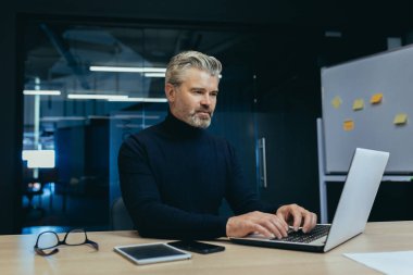 Modern ofisin içinde çalışan ciddi, olgun, gri saçlı iş adamı, iş yerinde laptopunu düşünceli bir şekilde daktilo eden adam..