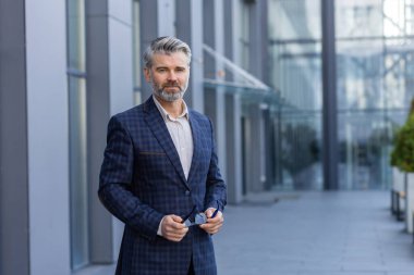 Portrait of successful mature man outside office building, businessman in business suit looking at camera thinking smiling while standing.