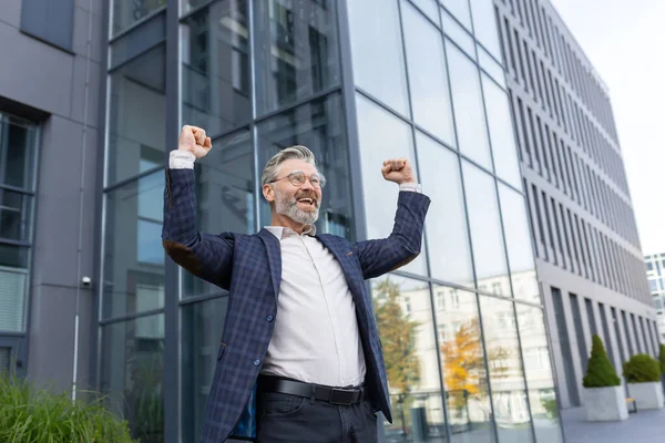 stock image Successful and happy gray-haired boss outside office building rejoices success and victory, triumphs with hands up, businessman business owner in business suit and glasses, mature investor banker