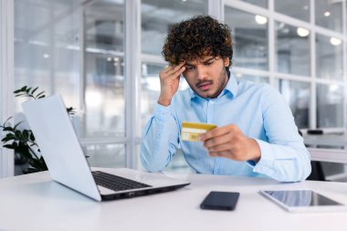 Hesapla ilgili problemler, iflas vs. Genç bir İspanyol adam ofisteki masada oturuyor, kredi kartına endişeli bir şekilde bakıyor, kafasını tutuyor..