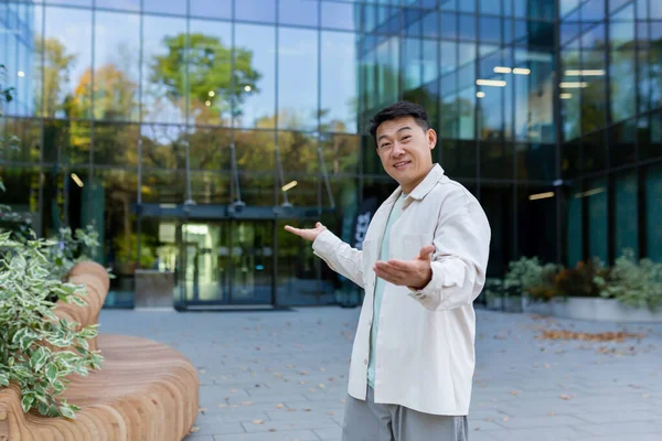 A young Asian man, businessman, company founder, director in casual clothes stands on the street outside and gestures to the camera to enter a modern office center, shows his workplace.