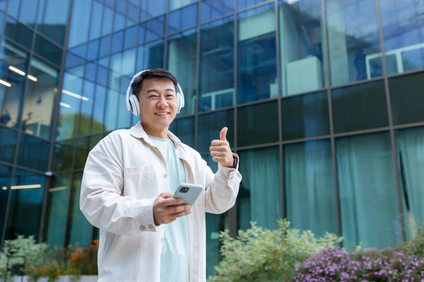 A young male Asian student stands outside the campus, co-working space. He listens to music, a podcast, an audiobook in white headphones from the phone, points a super finger at the camera, smiles.