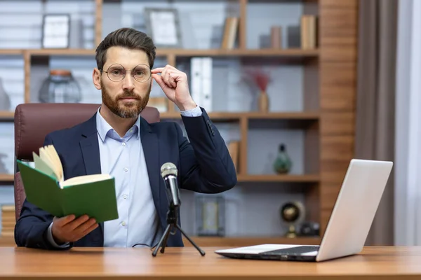 Online presentation of the book. Record a podcast. A young man writer, author, translator in a suit sits in the office at a table in front of a camera, a laptop and a microphone, holds a book.