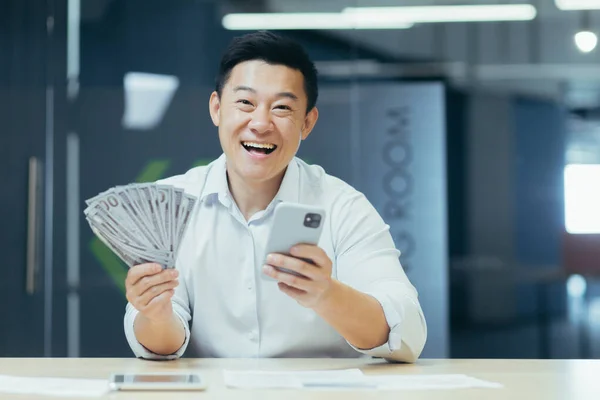A happy young Asian man sits in the office at the table, holds a phone and cash money in his hand. A smiling man dials the phone, rejoices at a monetary win, lottery.