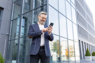 Mutlu kıdemli gri saçlı işadamı, avukat, yatırımcı ofis dışında duruyor, gökdelen. Neşeli telefon kullanır, okur, gönderir, alır..