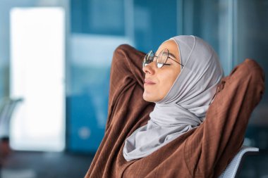 Yan taraftaki yakın plan fotoğraf. Yorgun, tesettürlü ve gözlüklü genç bir Arap kadın elleri başının arkasında bir sandalyede oturur, dinlenir, uyur, gözlerini kapatır..