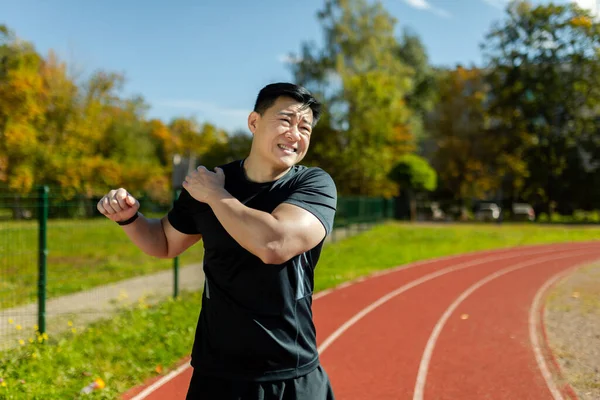 stock image Asian sportsman stretches his shoulder, man in the stadium after running and active physical exercises stretches his arm joints, has shoulder pain, arm muscle spasm.