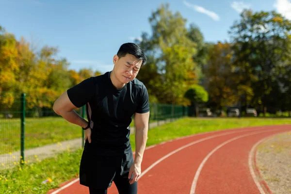 Asian sportsman has severe pain in back and abdominal muscles, man peed training in stadium on sunny day massaging side of back, athlete on jogging pulled muscle.