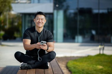 Genç bir Asyalı sporcu gölün yakınındaki ahşap bir bankta dinleniyor ve elinde kulaklık olan bir cep telefonu kullanıyor. Kameraya gülümsüyor..