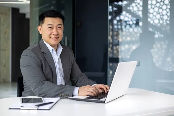 Successful mature asian boss working on laptop, portrait of adult senior investor at computer at workplace, businessman smiling and looking at camera, man in business suit typing on keyboard.