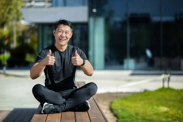 Asian young athlete, trainer, practitioner sits on the lake shore on a bench in the lotus position, meditates, does yoga. Smiling at the camera, he shows his fingers super, success.