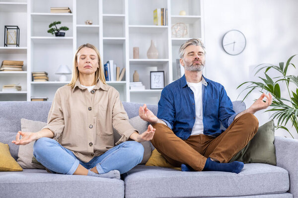 Adult mature couple man and woman relaxing together at home sitting on sofa in lotus position, family meditating together in living room.