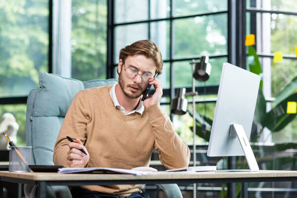 Communication with the client, partner. Young male office worker, businessman talking on the phone in the office, making notes with a pen.