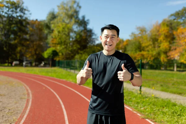 stock image A young Asian athlete, a coach stands in a stadium in a black sports uniform. Promotes sports, looks into the camera, shows super with fingers.