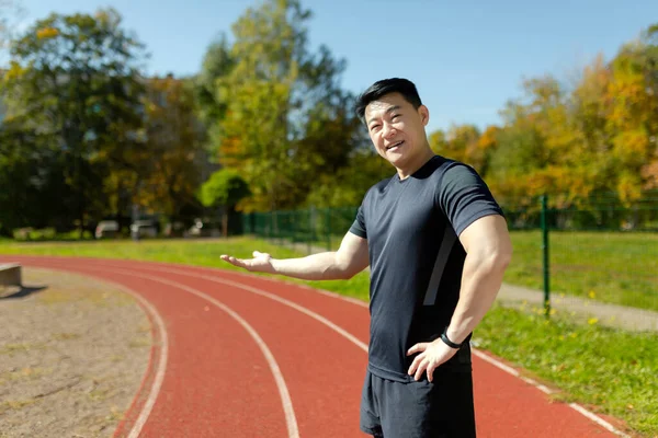 Young Asian athlete, coach standing in stadium and pointing with hands to running track. Invites to training, presents and popularizes sports