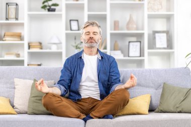 Yetişkin bir adam evde yalnız başına, Lotus pozisyonunda oturuyor, koltukta oturuyor, gri saçlı biri oturma odasında günlük kıyafetlerle meditasyon yapıyor..