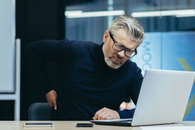 Sick senior man working inside office modern building, overtired mature businessman having severe back pain, boss massaging side to back, man at work with laptop.