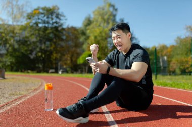 Genç bir Asyalı adamın portresi, koşu bandında telefonla oturan bir atlet. Sevinçliyim, zafer jesti yapıyorum, evet. Koşu, antrenman ve kutlama sonuçlarını kontrol eder.