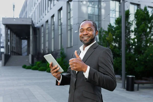 Portrait of a smiling and successful black senior businessman standing near an office center and working on a tablet. Looks at the camera and points a super finger.