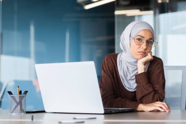 Başörtüsü içindeki üzgün kadın kameraya bakıyor, iş yerindeki iş kadını başarı sonuçlarından memnun değil, ofiste laptopla çalışan Müslüman kadını üzüyor..