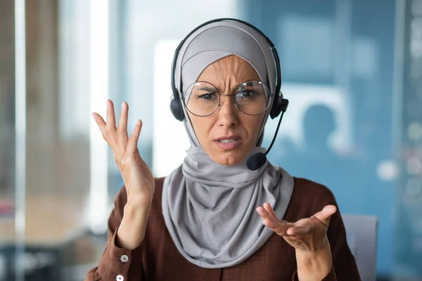 Angry woman in hijab talking on video call, businesswoman with headset phone close-up looking at web camera and talking to colleagues in online meeting.