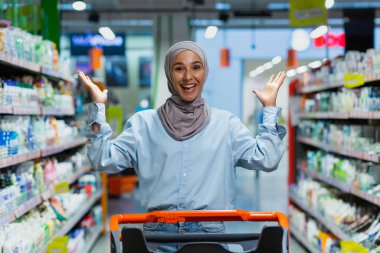 Evdeki kimyasallar bölümündeki bir süpermarkette alışveriş arabasıyla duran tesettürlü genç Müslüman bir kadının portresi. Elleriyle gösteriyor, kameraya bakıyor, poz veriyor, reklamlar yapıyor..