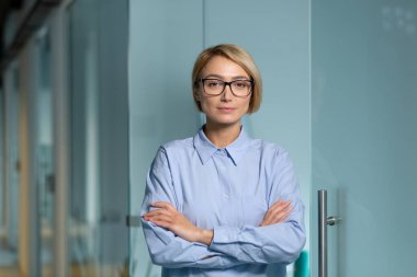Ciddi ve başarılı bir iş kadını iş yerindeki ofis patronu, kameraya dikkatlice bakan kadın çapraz kollu ve mavi gömlekli..