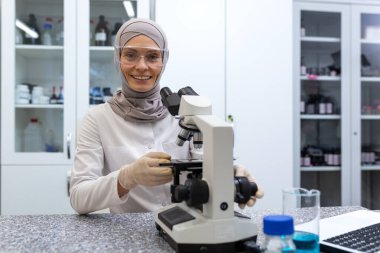 Genç Müslüman bir laboratuvar asistanının portresi, tıbbi teşhis merkezinde mikroskopla masada oturan bir doktor. Gülümseyerek kameraya bakıyor..