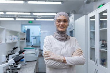 Genç Müslüman bir laboratuar teknisyeninin portresi tesettür ve koruyucu gözlüklü eczacı, tıbbi laboratuvarda duruyor, kolları göğsünde çaprazlanmış, kameraya gülümsüyor..