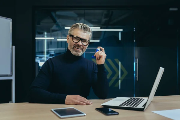 stock image A gray-haired senior man in glasses sits at a desk in an office and uses an inhaler. Looks into the camera.