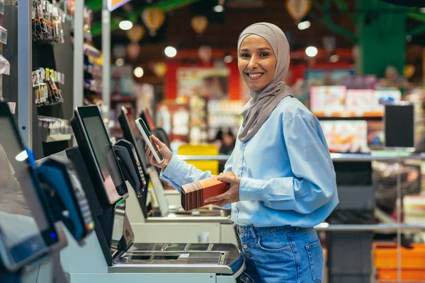 Self-service in a supermarket. A young Arab woman in a hijab is standing near the electronic cash registers and is paying for the goods in the store online through an application on her phone.