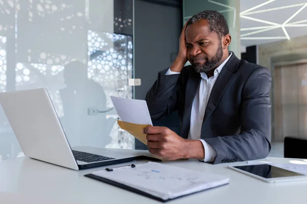 Nervous and upset businessman in office at workplace received mail envelope notification message with bad news bankruptcy, african american man sad and pensive reading document.