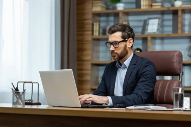 Successful mature businessman working inside office, man in business suit working at workplace with documents and laptop, boss behind paperwork attentive and focused.