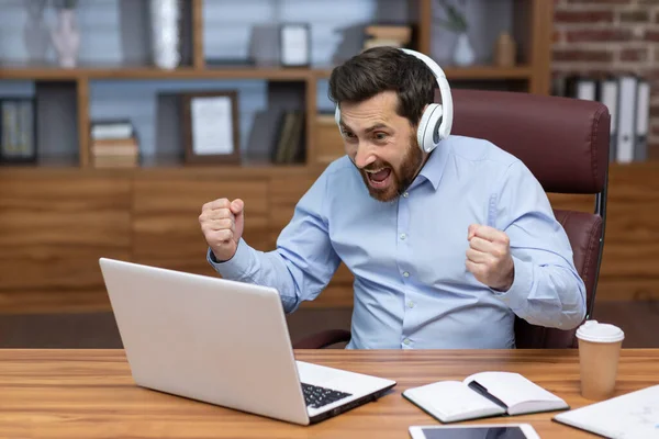 stock image Mature businessman in shirt and headphones watching sports match at workplace inside office, man cheering having fun at work, watching video on laptop.