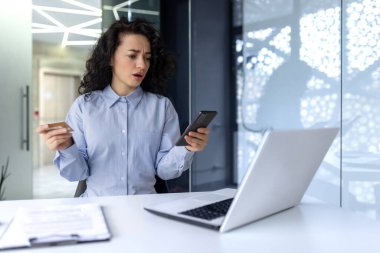 Kredi kartı sorunları. Genç bir Arap kadın ofiste masada oturuyor ve endişeli bir şekilde telefona bakıyor. Başarısız alışveriş, engellenmiş kanal.