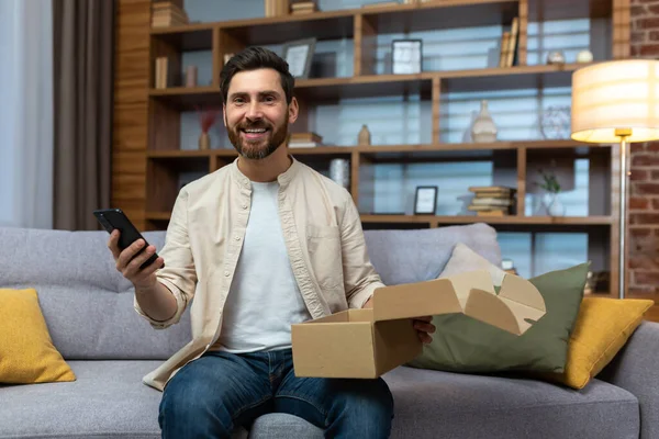 Portrait of happy online shopper at home, mature adult man smiling and looking at camera, holding product box and phone, happy about fast delivery from online store.