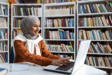 Türbanlı genç ve güzel bir öğrenci üniversite kütüphanesinde uzaktan öğrenim görüyor, dizüstü bilgisayarında yazı yazıyor bilgi arıyor ve sınav için hazırlanıyor..