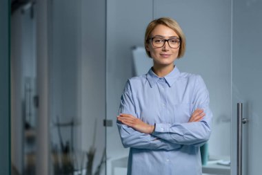 Genç, güzel bir kadın tasarımcının portresi, mimar, programcı kollarını göğsünde kavuşturarak ofiste duruyor. Kendine güvenerek ve gülümseyerek kameraya bakarak..