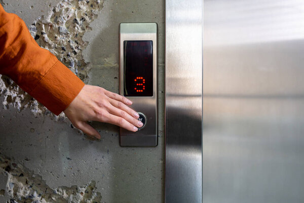 Close-up photo. The hand of a young woman in an orange shirt pressing a metal elevator button with her finger in an office center, hotel, house.