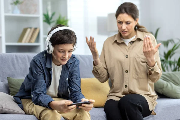 Family conflict quarrel, woman mother quarrels with son, teenage boy in headphones ignores woman and plays video games on phone, family at home in living room sitting on sofa.