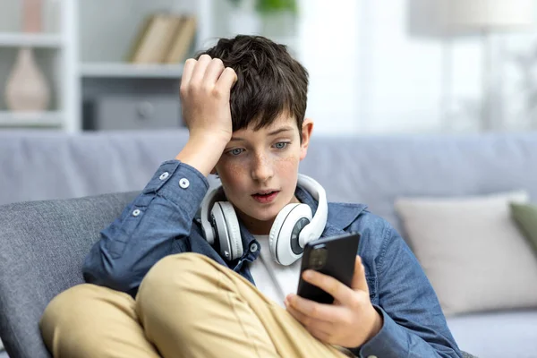 Young teenager boy reading bad news on phone, son sitting on sofa in living room with headphones at home closeup.