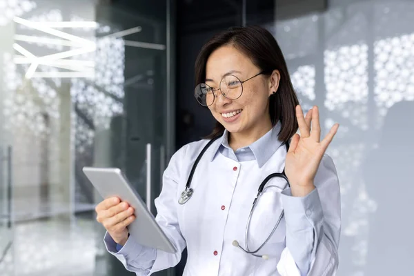 快乐且面带微笑的亚洲女医生与病人进行远程对话 妇女站在诊所 使用平板电脑进行视频通话和在线病人咨询 — 图库照片