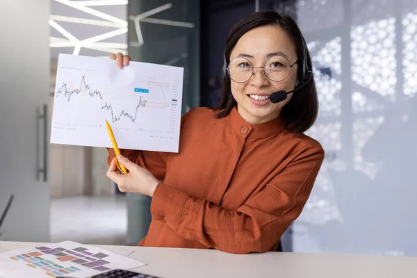 Online meeting video meeting discussion, Asian woman looking at camera and showing forex chart, businesswoman smiling and telling financial business project presentation, webcam view.