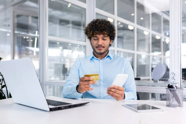 Stock image A young male student is sitting at a desk on campus and holding a phone and a credit card. Pays the bill for education, housing rent, credit loan.